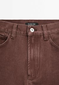Pantalon en denim marron avec fermeture à boutons sur le devant, passants pour ceinture, poches avant et une étiquette Massimo Dutti visible à l'intérieur de la ceinture.