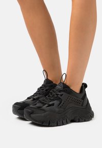 Sneaker sportive nere con una toma superiore testurizzata, caratterizzate da dettagli in rete, finiture perforate, una suola spessa e modelli di battistrada prominenti.
