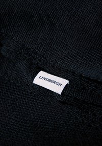 Zwart gestructureerde stof met een geweven patroon, met een wit label waarop "LINDBERGH" in marineblauw is gedrukt. Close-up perspectief.