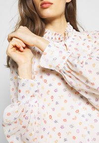 Blouse blanche à manches longues avec un col smocké, ornée d'un motif floral en violet, orange et jaune. Tissu texturé avec des manches bouffantes.