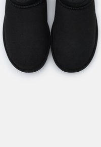 Paire de chaussures noires sans lacets, avec des bouts arrondis et des dessus en tissu doux, sur un fond gris neutre.