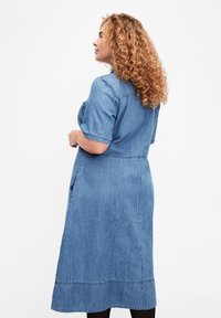 Denimmekko lyhyillä hihoilla, keskipitkä ja suora leikkaus. Siinä on sivutaskut ja näkyvät saumat helmassa. Vaaleansininen väri.