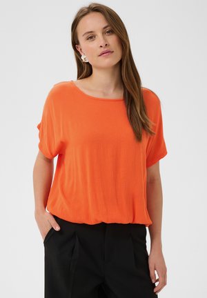 Femme aux longs cheveux bruns portant un haut orange à manches courtes et un pantalon noir, une main dans la poche, fond neutre.