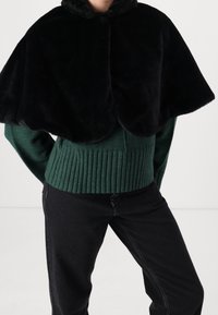 Svart falsk pälscape med rundade kanter, bärs över en mörkgrön ribbad tröja. Modellen står med händerna på höfterna och har på sig svarta jeans.