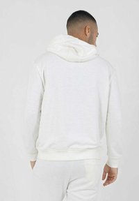 Sudadera blanca con capucha y una textura suave, que cuenta con un bolsillo tipo canguro, puños acanalados y un ajuste relajado. Detalles de costura visibles.