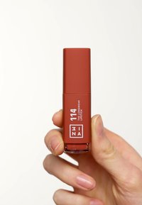 Brown lipstick tube with matte finish, white text label, and clear base. Held by a hand against a light background.