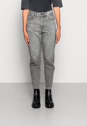 Personne portant un jean gris délavé taille haute et des bottines noires avec fermeture éclair argentée et anneau, debout devant un fond uni.