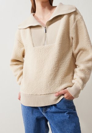 Person trägt einen beigen Sherpa-Fleece-Pullover mit Frontreißverschluss und geripptem Kragen, Bündchen und Saum, kombiniert mit blauen Denim-Jeans.