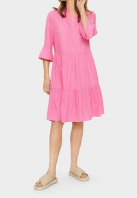 Rosa Stufenkleid mit V-Ausschnitt und dreiviertellangen Ärmeln. Verfügt über geraffte Ärmelbündchen und eine lockere Passform. Kombiniert mit beigen Slip-On-Sandalen.