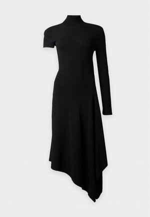 Femme aux longs cheveux foncés portant une robe noire asymétrique avec une manche courte et une manche longue, associée à des sandales noires à talons hauts.