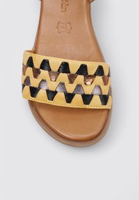 Sandale en cuir jaune à enfiler avec une large bande ornée de motifs en zigzag noir, bronze et argent, reposant sur une semelle lisse marron.