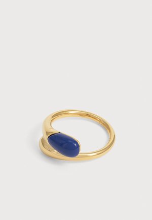 Bague ouverte en or avec une pierre lisse en forme de goutte d’eau bleu foncé sertie à une extrémité sur un fond clair uni.