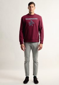 Sudadera burdeos con "VALCUATRO" impreso en negro, escote redondo, puños de canalé, combinada con jeans ajustados gris claro y zapatos marrones.