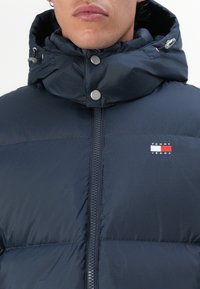 Tamno plava puffer jakna s visokim ovratnikom, zatvaranjem na patentni zatvarač i gumbima; sadrži Tommy Jeans logo na prsima; glatka, quilted tekstura.