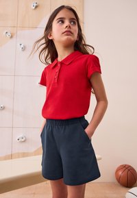 Rotes Baumwoll-Polohemd mit Kragen und drei Knöpfen, kombiniert mit marineblauen Shorts mit elastischem Bund und seitlichen Taschen.