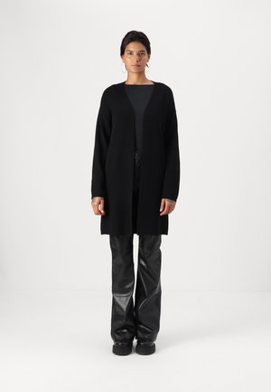 Melna garā kardigāna no mīkstas adīšanas materiāla, ar atvērtu priekšpusi bez aizdariem, savienots ar melnām platiem ādas biksēm un masīviem melniem zābakiem.