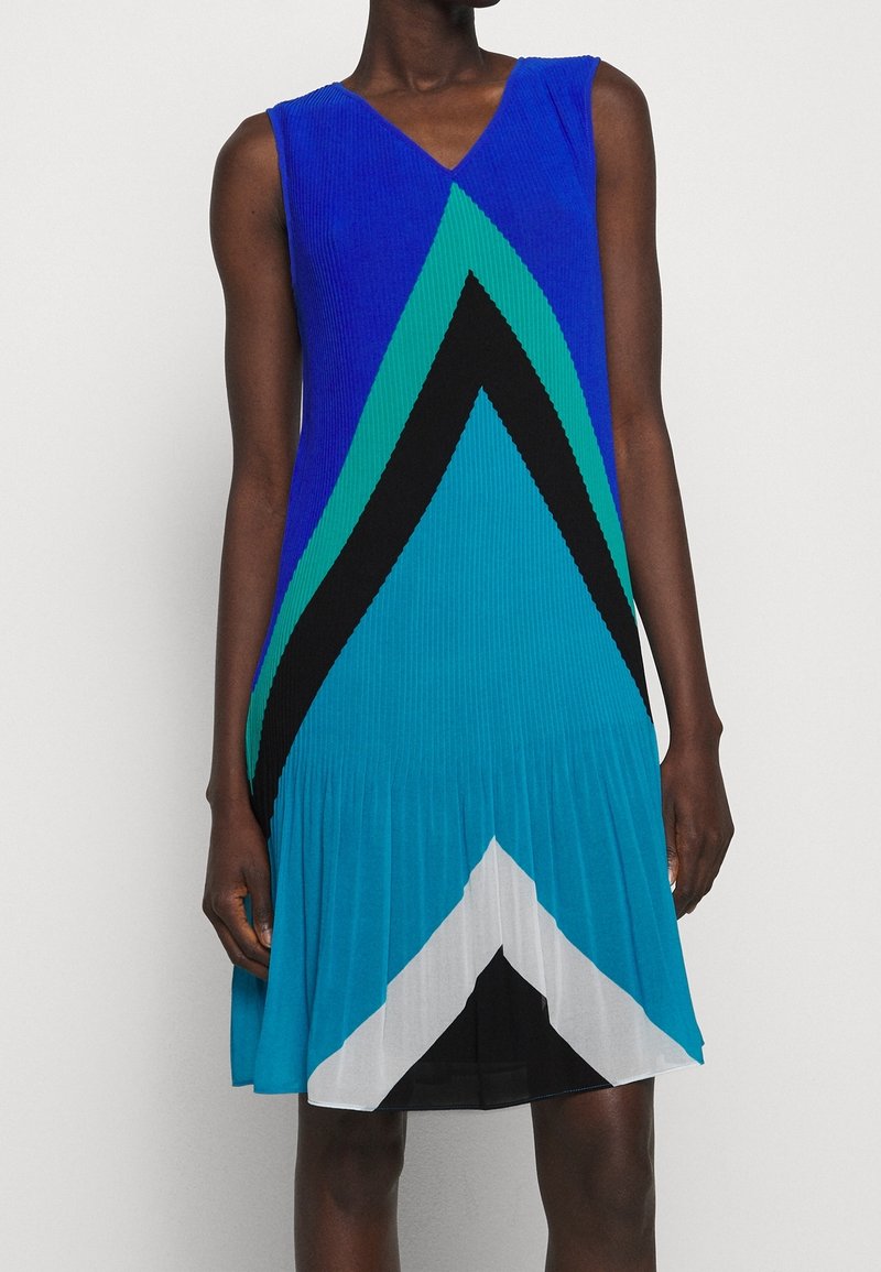 Robe en maille avec un col en V, présentant des motifs géométriques audacieux en bleu, vert, noir et blanc. La jupe plissée évasée ajoute de la texture et du mouvement.