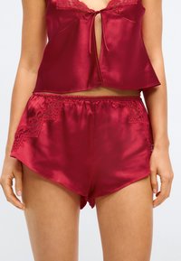 Rød sateng nattøysett som inkluderer en kort topp med knyting og blondeaccenter, kombinert med matchende shorts med blonde-detaljer på sidene.