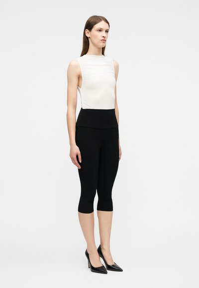 Γυναίκα όρθια, φορώντας λευκή αμάνικη μπλούζα, μαύρο κοντό ελαστικό παντελόνι και μαύρα παπούτσια με ψηλό τακούνι, μπροστά σε απλό γκρι φόντο.