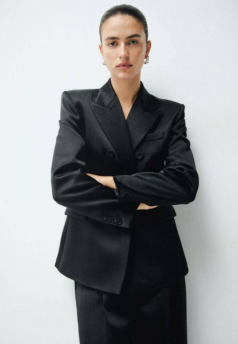 Blazer negro ajustado con solapas de pico, doble botón y bolsillos frontales. Textura suave, combinado con pantalones a juego de cintura alta.