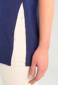 Mörkblå t-shirt med sidoflikar, tillverkad av mjukt material. Har en avslappnad passform och bärs över ljusa byxor. En hand synlig från sidan.