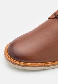 Scarpa in pelle marrone con punta arrotondata, caratterizzata da cuciture arancioni lungo il bordo e una suola in gomma beige. Texture liscia, design minimalista.