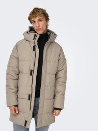 Beige puffer coat with a hood, featuring a quilted pattern, front zipper, and contrasting black hardware. Worn over a black turtleneck.