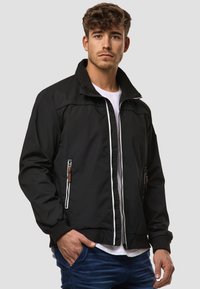 Black bomber jacket with a high collar, zippered pockets, and textured fabric. Features white zipper accents and a fitted waist.