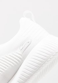 Vita stickade slip-on sneakers med strukturerad sula och varumärkeslogga på sidan, med en dragflik vid hälen.