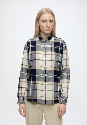 Donna con capelli lisci biondi indossa una camicia a quadri beige e blu navy con bottoni e pantaloni beige, in piedi davanti a uno sfondo bianco semplice.