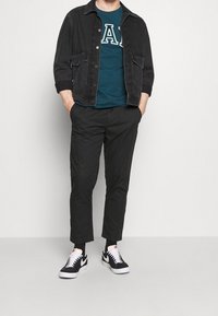 Zwarte spijkerjack over een teal graphic t-shirt met de letters "A" en "A", gecombineerd met zwarte tapse broek en zwarte Nike sneakers.