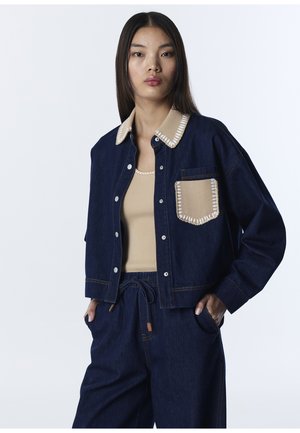 Jonge vrouw die een donkerblauw spijkerjack draagt met een beige gestikte kraag en zak, bijpassende wijde broek en een beige geribd tanktopje.