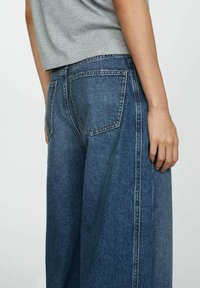 Široke denimske hlače v temno modri barvi z gladko teksturo. Imajo velike zadnje žepe in kontrastne šive ob šivih.
