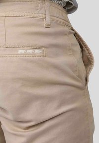 Pantalon kaki en mélange de coton ; présente une poche arrière avec trois silhouettes de vaches blanches et des détails de couture subtils. Texture lisse.