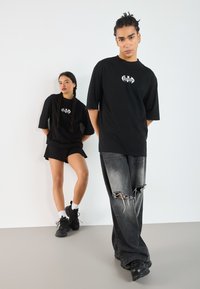 Black oversized t-shirts with graphic logos, paired with distressed black jeans and black sneakers. Smooth fabric with loose fit design.