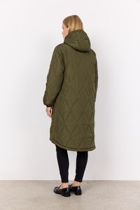 Vrouw in een lange olijfgroene doorgestikte gewatteerde jas, zwarte slim fit broek en zwarte instappers, staand in zijaanzicht op een effen achtergrond.