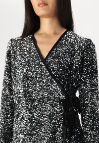 Μαύρο φόρεμα σε βελούδο με στρας, μακρυά μανίκια, βαθύ V ντεκολτέ και ζώνη που δένει μόνη της. Διαθέτει υφή από πυκνά τοποθετημένα στρας.