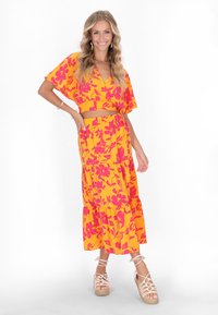 Setti crop top ja maxi hame kirkkaan keltaisena, jossa on vaaleanpunaisia kukkakuvioita. Kangas on kevyt; hameessa on kerroksellisia röyhelöitä. Yhdistetty espadrille-sandaalien kanssa.