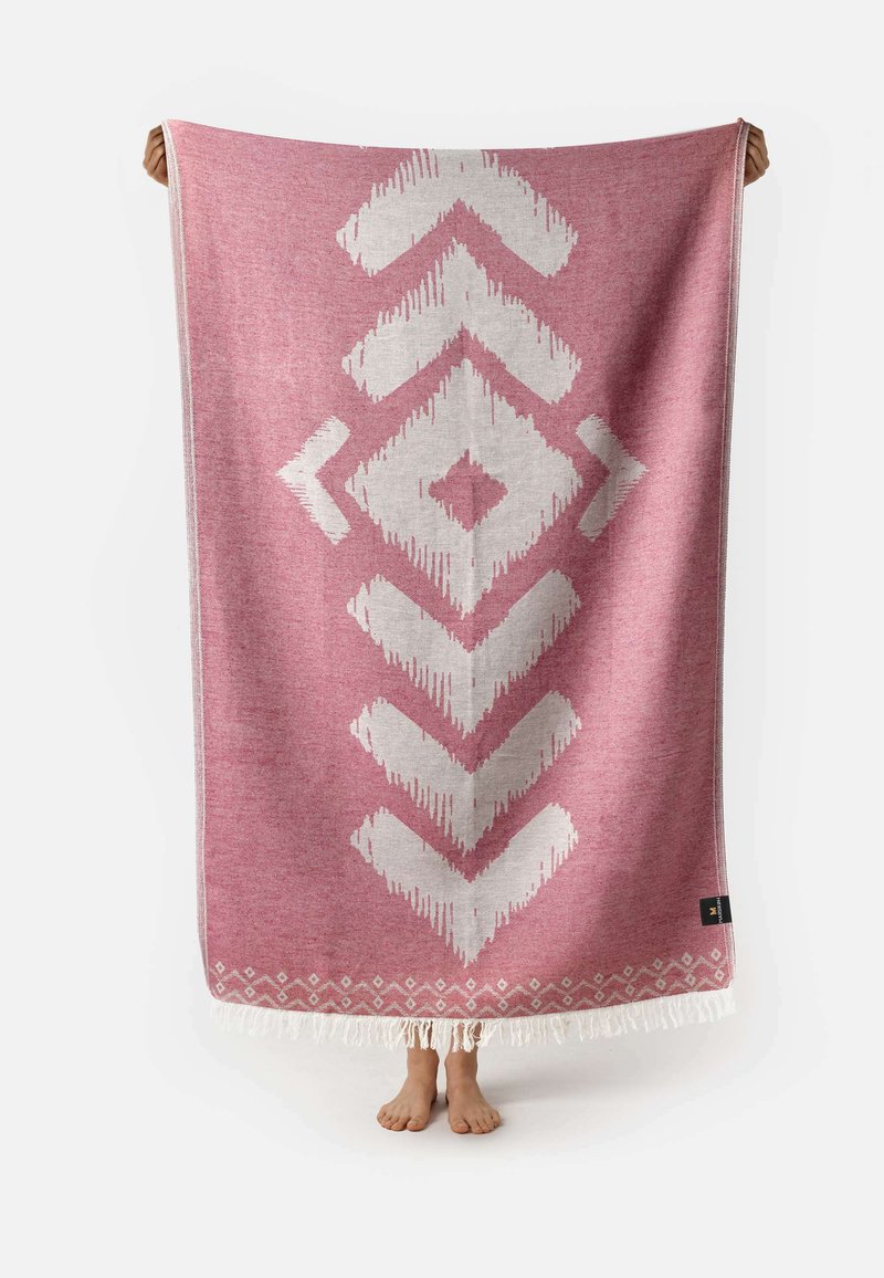 Manta rectangular de color rosa con un patrón geométrico blanco. Fabricada en tela suave, cuenta con bordes con flecos. El patrón incluye diamantes y flechas.