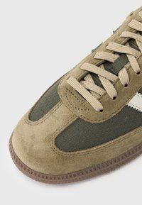 Oliivinvihreä tennari, joka on tehty mokkanahasta ja teksturoidusta kankaasta. Siinä on beiget nauhat, ruskea kumipohja ja valkoiset sivukoristelut.