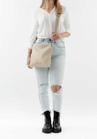 Borsa a tracolla in pelle beige con una tracolla testurizzata, forma rettangolare, accessori in argento, indossata da una persona con jeans strappati azzurri chiari e stivali neri.