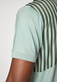 Maglietta sportiva verde chiaro a maniche corte con un motivo a strisce azzurro scuro sulla parte posteriore. Tessuto liscio con un design aderente.