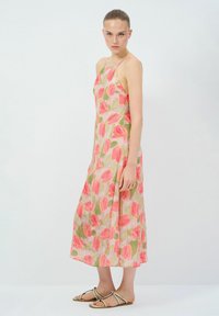 Bloemige midi-jurk van een lichte stof met roze en groene tulpentjes, dunne bandjes en een ontspannen silhouet. Model draagt platte sandalen.