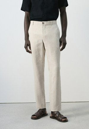 Persona che indossa pantaloni beige a gamba dritta, camicia nera a maniche corte e sandali in pelle marrone scuro, in piedi su pavimento grigio contro una parete bianca.