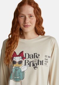 Ruda kobieta w beżowej bluzie, na której znajduje się postać Daisy Duck w okularach przeciwsłonecznych, z czerwoną kokardą oraz napisem "Odważ się być jasna".