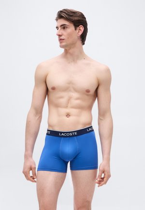 Άνδρας νέος, καλογυμνασμένος, χωρίς πουκάμισο, με κοντά καστανά μαλλιά, φοράει μπλε μποξεράκι Lacoste, στέκεται μπροστά σε απλό φωτεινό φόντο.