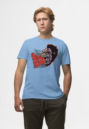 Ung mand iført en lyseblå t-shirt med en farverig grafik af et brølende gorillahoved og "Skull Island" tekst i rødt.