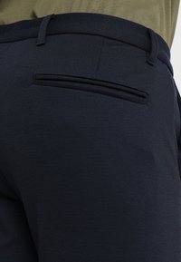 Gros plan sur un pantalon sur mesure bleu marine foncé avec une poche passepoilée à l'arrière et des passants pour ceinture, porté avec un haut vert olive.