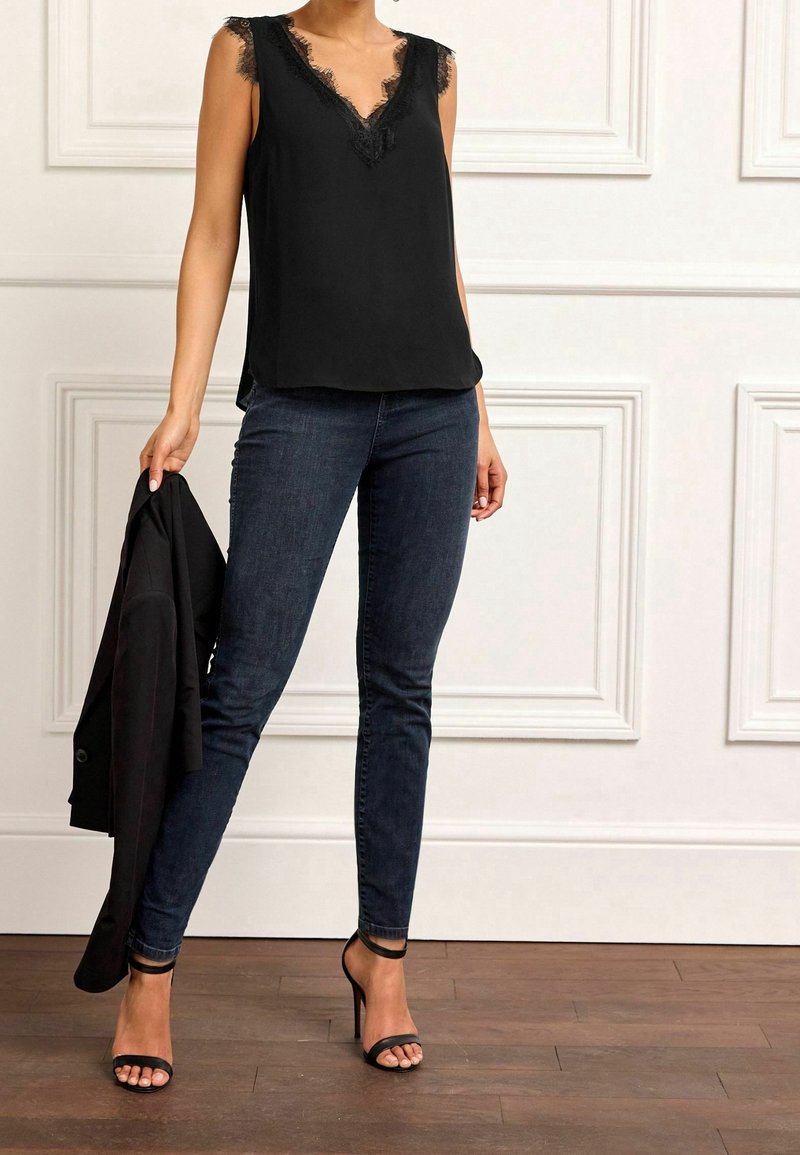 Haut noir sans manches avec bordure en dentelle au niveau du décolleté, associé à un jean foncé et des escarpins noirs à brides. Veste tenue dans une main.