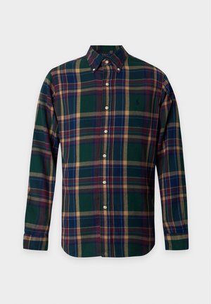 Homme debout portant une chemise à carreaux verte et bleue boutonnée, un pantalon jaune moutarde et des baskets blanches sur fond blanc.